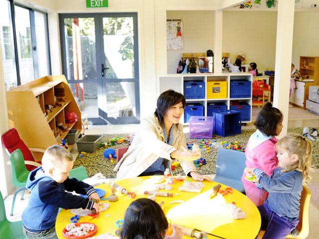 Children playing at table with teacher with floor play area in background.